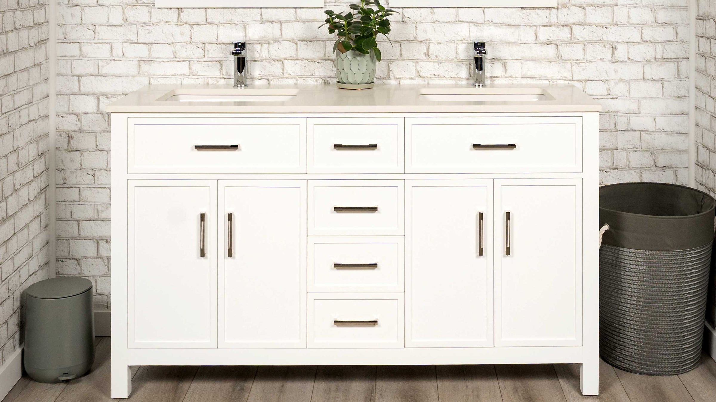 A white double-sink bathroom vanity cabinet with six drawers and two cabinet doors, featuring slim shaker design. The vanity is set against a white brick wall, with a small potted plant on top, and a gray metal trash can on each side.