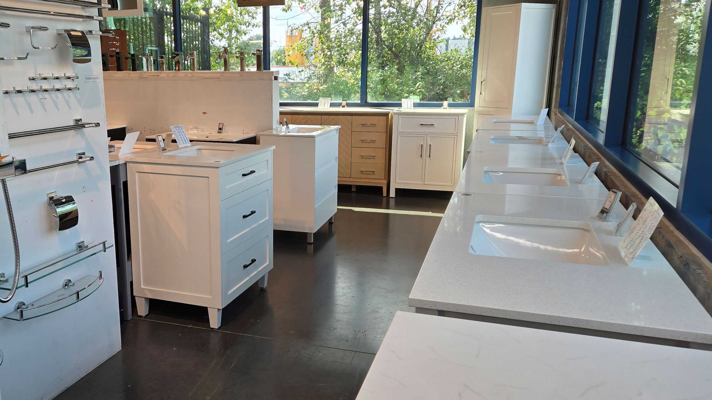A showroom interior with multiple white bathroom vanity cabinets, sinks, faucets, and wall-mounted fixtures arranged in a bright space with large windows and dark flooring.