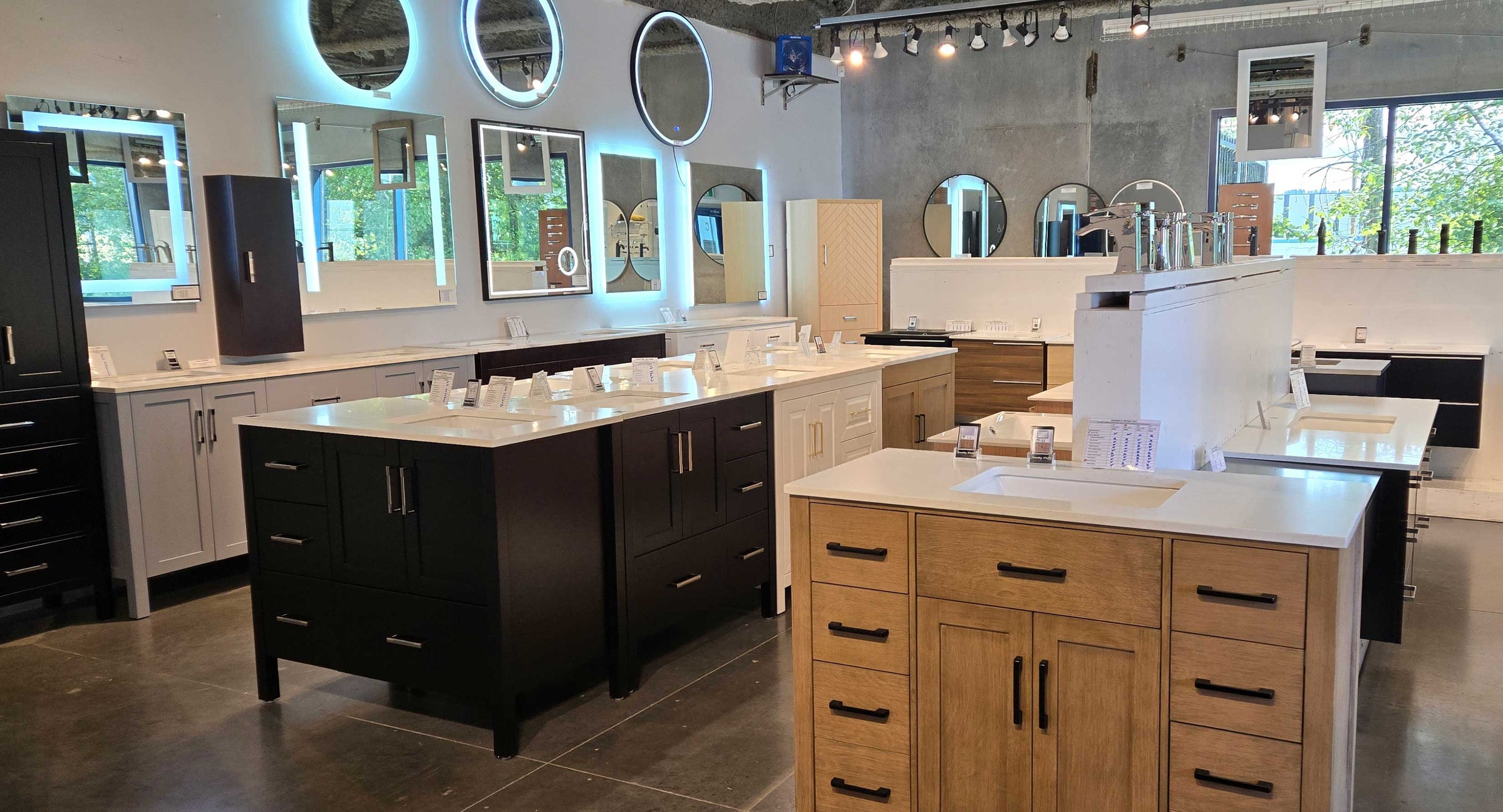A showroom interior displaying multiple bathroom vanity sets, mirrors, and faucets. In the foreground, a light wood vanity with a white countertop and integrated sink is visible, alongside other dark wood and light wood vanities with matching mirrors and fixtures.
